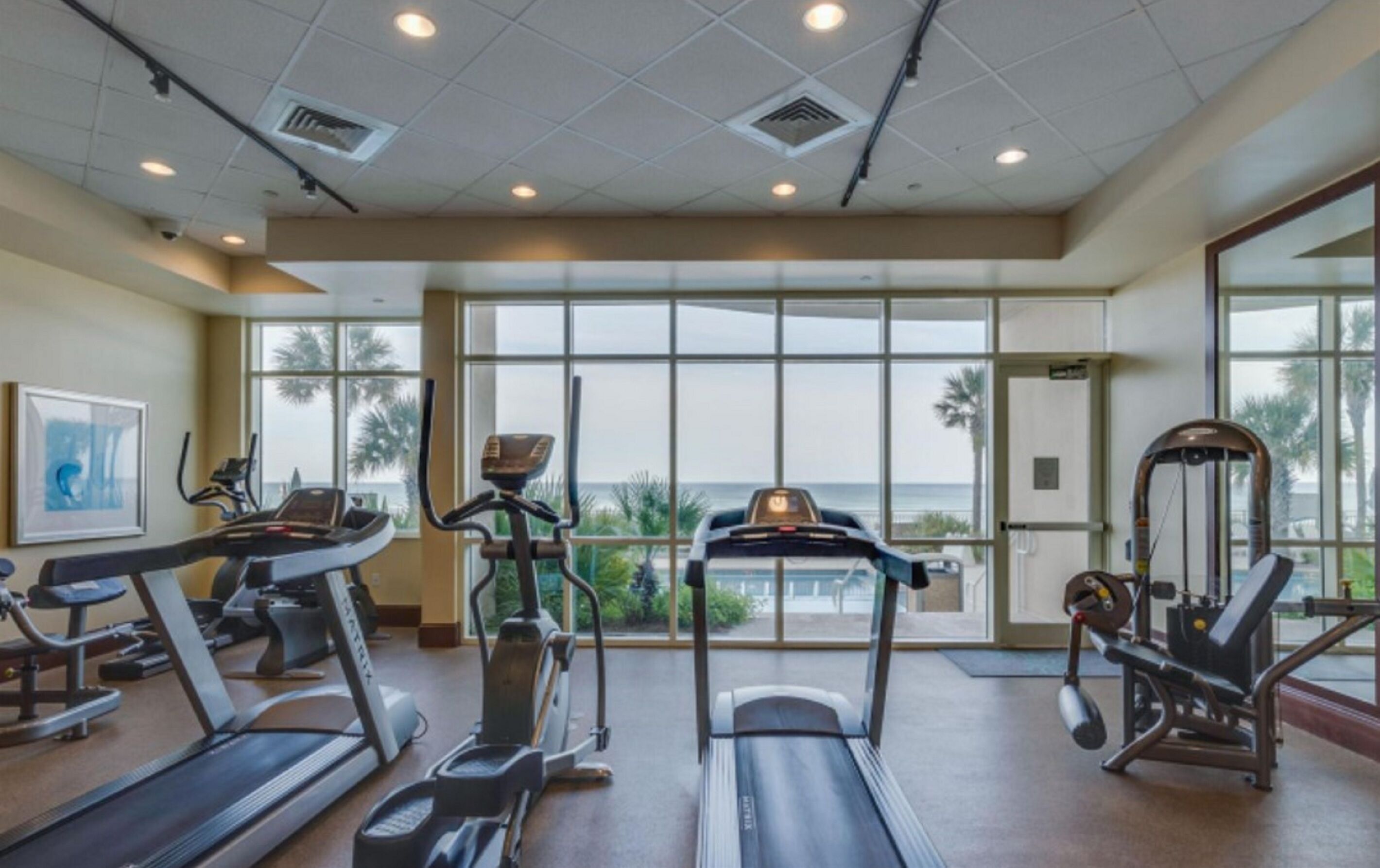 Une salle de sport équipée de tapis de course, de vélos elliptiques et d’appareils de musculation. De grandes fenêtres offrent une vue sur les palmiers et l’océan.