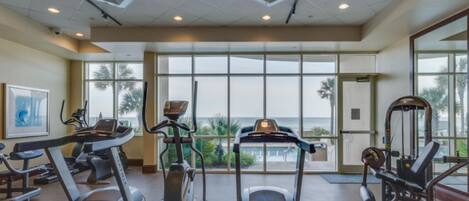 Une salle de sport équipée de tapis de course, de vélos elliptiques et d’appareils de musculation. De grandes fenêtres offrent une vue sur les palmiers et l’océan.
