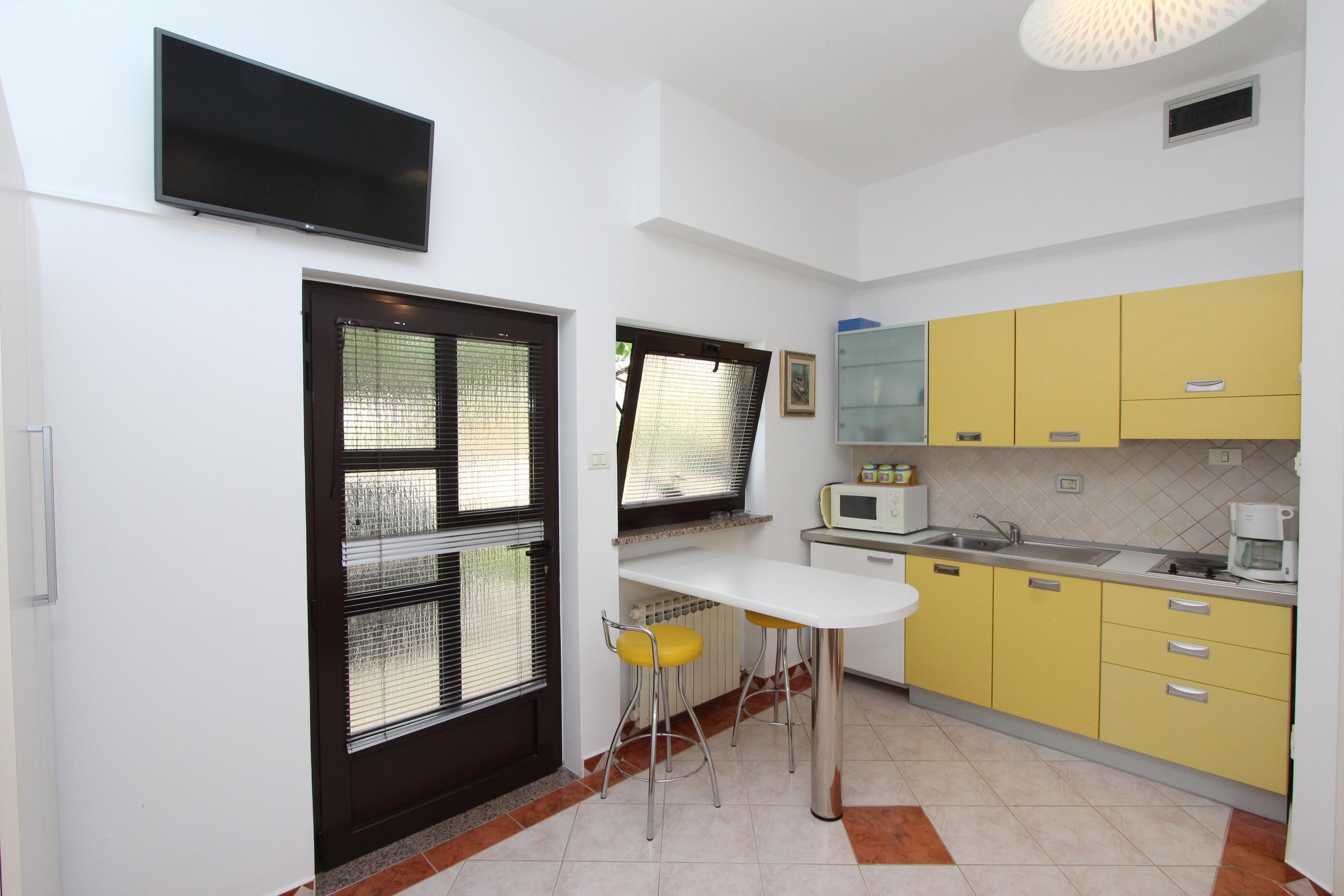 Cocina compacta con encimera blanca, armarios amarillos, ventana con marco negro y un televisor de pantalla plana sobre la encimera.