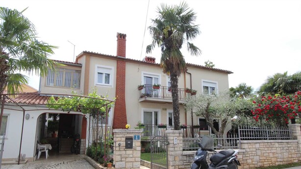 A two-story residential building with a stone entrance gate, a parked scooter, and a balcony with potted plants.