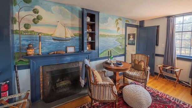 Een gezellige woonkamer met een open haard, een groot schilderij van een zeilboot op het water en uitzicht op de omgeving door een raam.