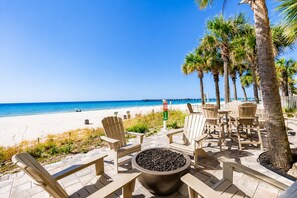 A beachside patio with Adirondack chairs, a fire pit, and palm trees.