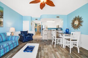 Un salón con un sofá azul, un sillón azul y una mesa de centro. Hay una zona de comedor con una mesa y sillas. Las paredes son azul claro y el suelo es de madera.