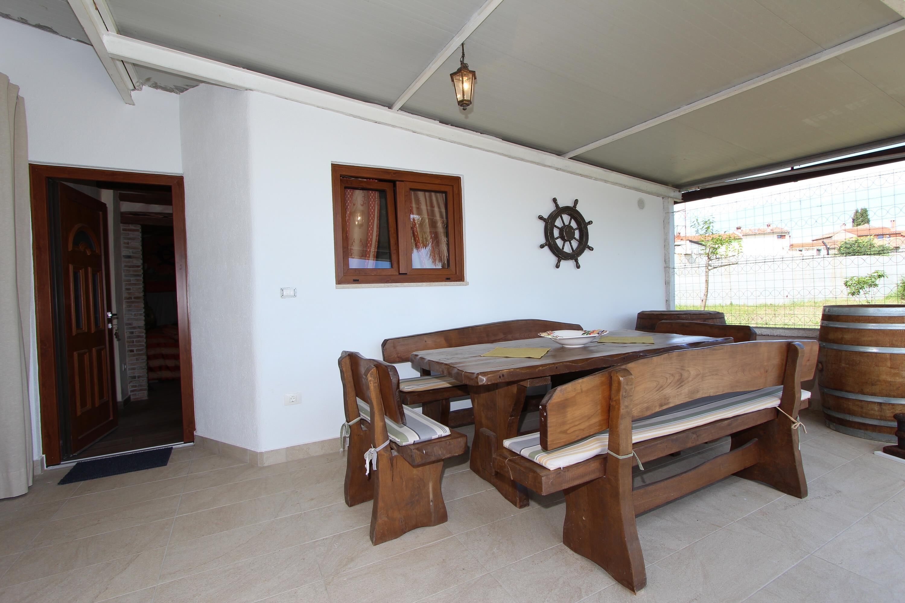 A dining area with a wooden table and benches, a ship's wheel decoration, and a view of the outside through a large window.