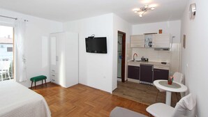 Una habitación de hotel pequeña con una cama, un banco verde, un televisor, una kitchenette y un baño visible a través de una puerta abierta.