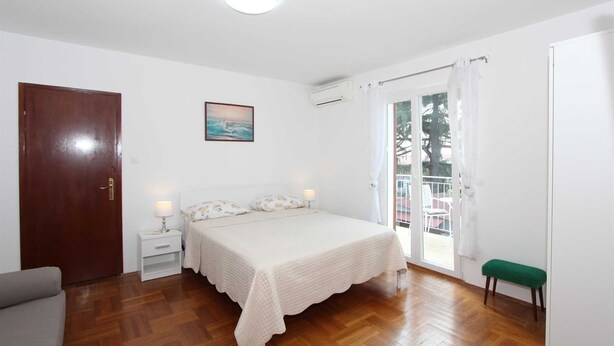 Un dormitorio con cama, mesita de noche, un banco verde, una puerta y una ventana con cortinas.