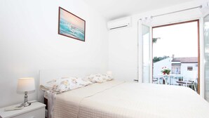 Un dormitorio con cama, mesita de noche con lámpara, un cuadro enmarcado en la pared y un equipo de aire acondicionado.