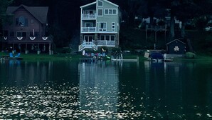 A large, multi-story house with a dock extending into a lake, surrounded by dense forest.