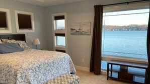 A bedroom with a large bed, a bench, a window with a view of the water, and a painting on the wall.