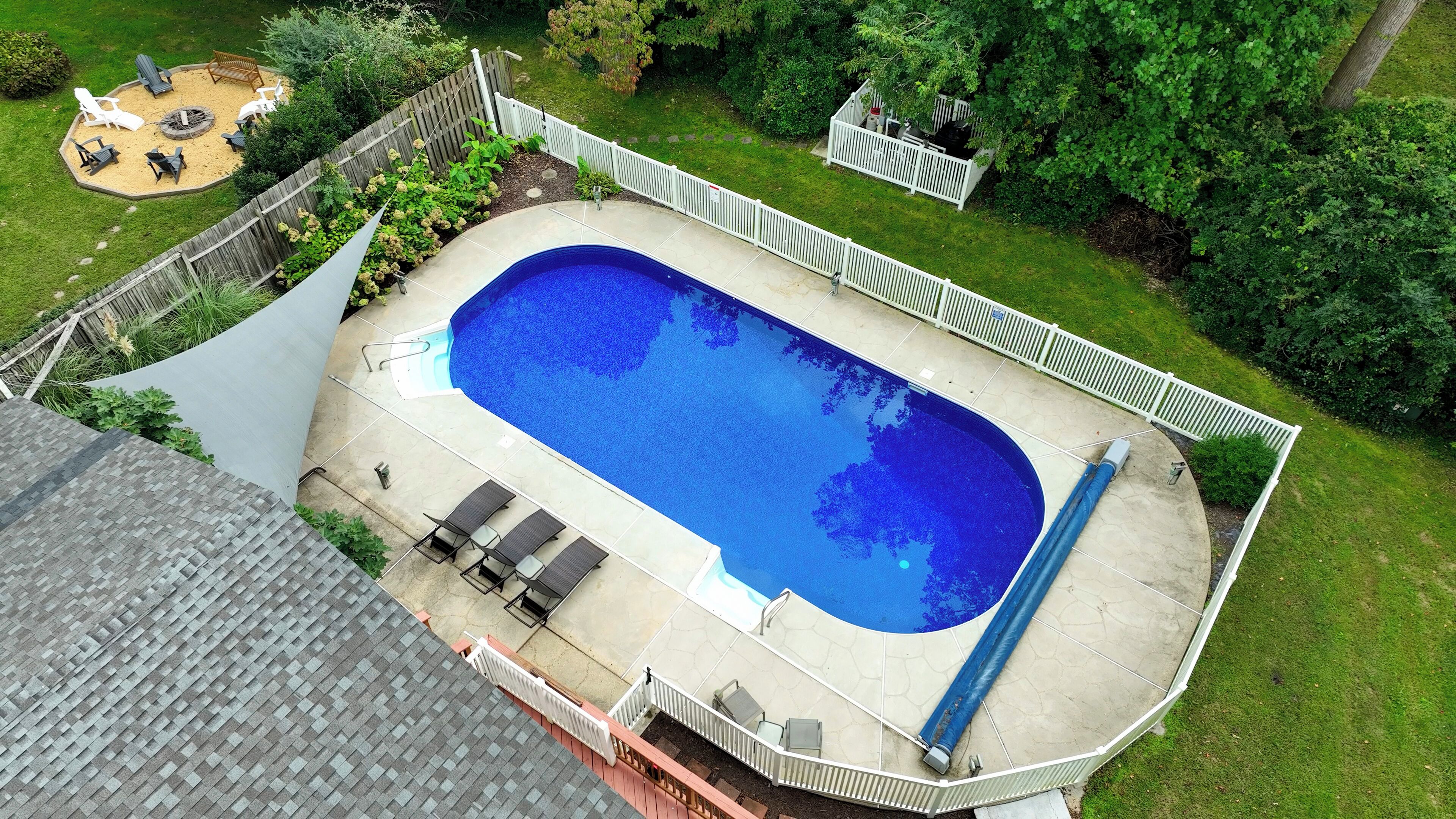 Une piscine rectangulaire dotée d’une plage blanche, entourée d’une clôture blanche, et un petit espace patio aménagé avec des chaises et un foyer.