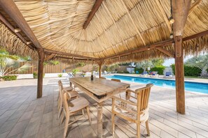 Une terrasse en bois surmontée d’un toit de chaume, une piscine et des chaises longues.
