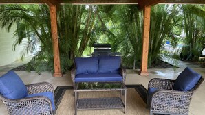 Een overdekt terras met rieten meubilair en een glazen salontafel, omgeven door weelderige begroeiing.