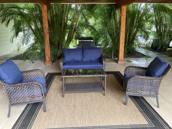 A covered outdoor seating area with wicker furniture and a glass-top coffee table, surrounded by lush greenery. - Tropical Guest House (Fort Pierce)