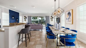 Uma cozinha moderna com uma ilha central, uma área de refeições com cadeiras azuis e uma sala de estar com sofá e televisão.