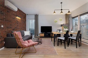 Ein modernes Wohnzimmer mit Backsteinwand, einem pinken Sessel, einer schwarzen Couch, einem Esstisch mit Stühlen, einem Fernseher und einem Fenster mit Jalousien.