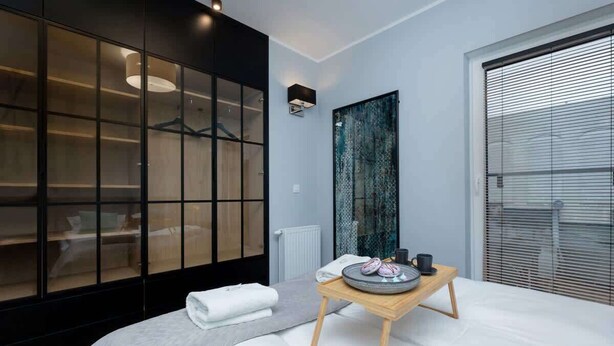 A modern hotel room with a large bed, a wooden side table, a wardrobe with glass doors, and a view through a window with blinds. - Elegant Apartment Near the Old Market Square | Parking (Poznań)