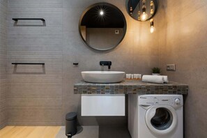 Un baño moderno con un espejo redondo grande, un lavabo, un perchero, una lavadora y una pequeña planta en maceta.