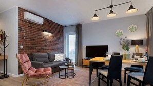 A modern living room with a brick wall, a sofa, a pink chair, a dining table with chairs, a TV, and a window with curtains. - Elegant Apartment Near the Old Market Square | Parking (Poznań)