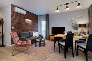 A modern living room with a brick wall, a sofa, a pink chair, a dining table with chairs, a TV, and a window with curtains. - Elegant Apartment Near the Old Market Square | Parking (Poznań)