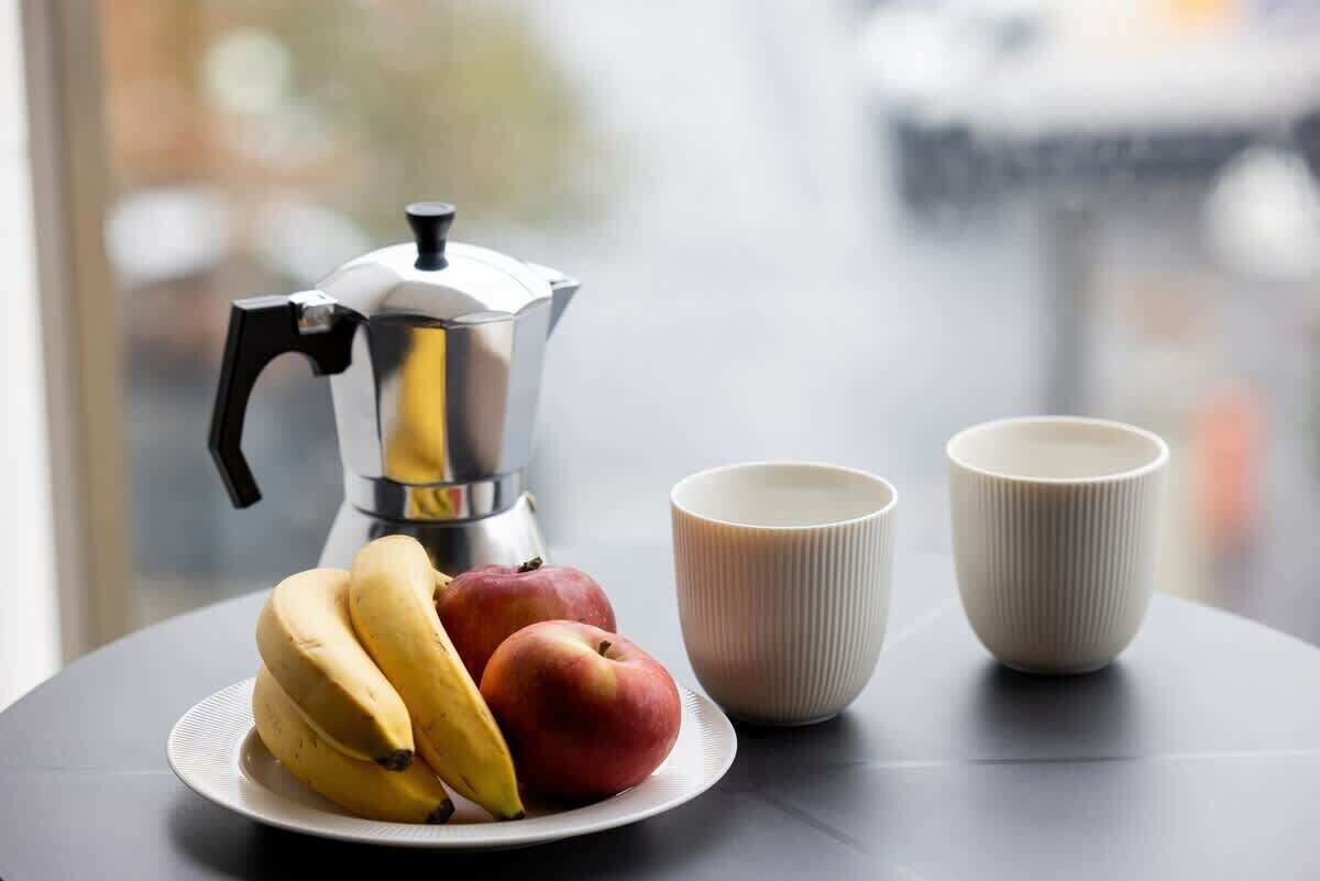 A table with a coffee maker, a plate of bananas and apples, and two cups.