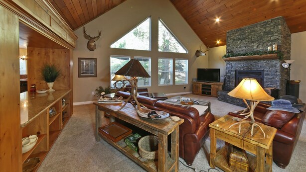 A spacious living room with a high ceiling, wooden beams, a stone fireplace, leather furniture, and large windows. - Comfortable Shaver Lake Getaway (Shaver Lake)