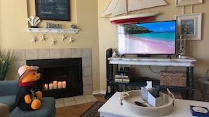 A living room with a fireplace, a television mounted on a white shelf, a plush toy on a blue sofa, and a nautical-themed decor including a sailboat model and starfish. - "The Erin" ~ a nautical condo with beach & pond view (Corpus Christi)