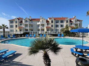 A pool area with blue lounge chairs and a picnic table, surrounded by a building with multiple balconies and a red roof.