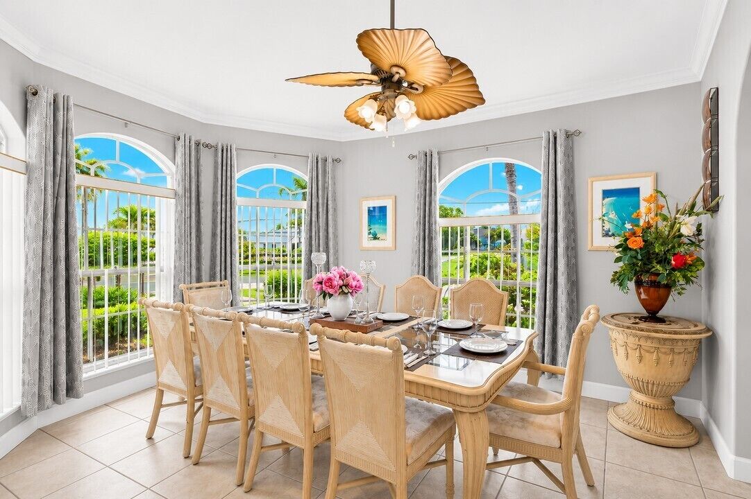 A dining area with a table set for four, surrounded by wicker chairs, with a view of greenery through large windows.