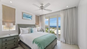 Une chambre moderne avec un grand lit, un ventilateur de plafond, une table de chevet, une lampe et une vue sur l’océan.