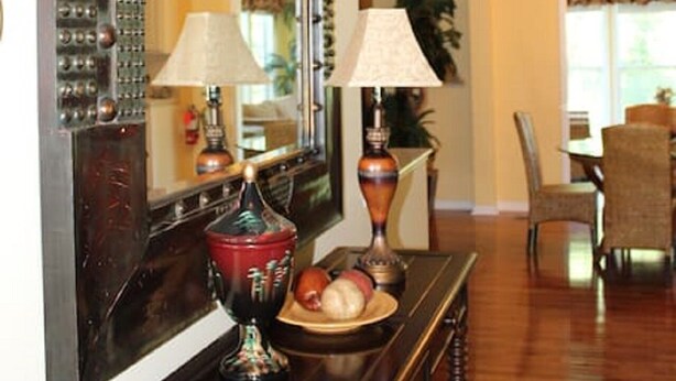 Een houten console tafel met een spiegel en twee lampen, een schaal fruit en een stoel in een eetgedeelte.