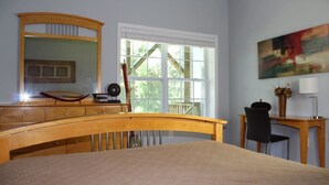 Een slaapkamer met een bed, een houten bureau, een stoel, een groot raam en een schilderij aan de muur.