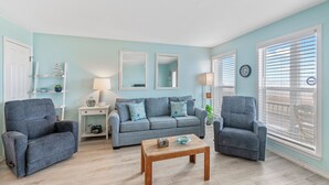 A living room with a light blue wall, a grey sofa with starfish pillows, two armchairs, a wooden coffee table, and a white door. - Gulf View Beachfront Escape | Pool, Hot Tub & Pet-Friendly (Corpus Christi)