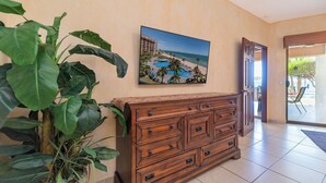 Una habitación de hotel con un aparador de madera, una planta grande en maceta y un cuadro de un complejo turístico en la playa.