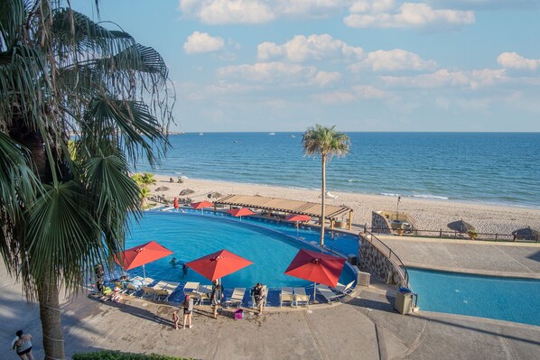 A pool area with red umbrellas, lounge chairs, and a view of the beach and ocean.