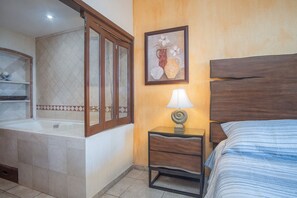 A bedroom with a bed, a nightstand with a lamp, a framed picture on the wall, and a bathroom with a bathtub and a shower.