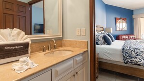 Un baño con lavamanos, espejo y caja de removedor de maquillaje. La habitación tiene una cama con ropa de cama azul y blanca, un sillón de cuero rojo y una ventana con cortinas.
