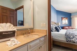 A bathroom with a sink, mirror, and makeup remover box. The room has a bed with blue and white bedding, a red leather chair, and a window with curtains.