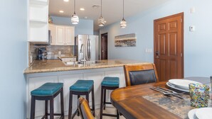 Cocina compacta con armarios blancos, un refrigerador de acero inoxidable y una encimera de granito. Hay tres taburetes de bar con cojines color turquesa y una mesa de comedor de madera con un mantel individual.
