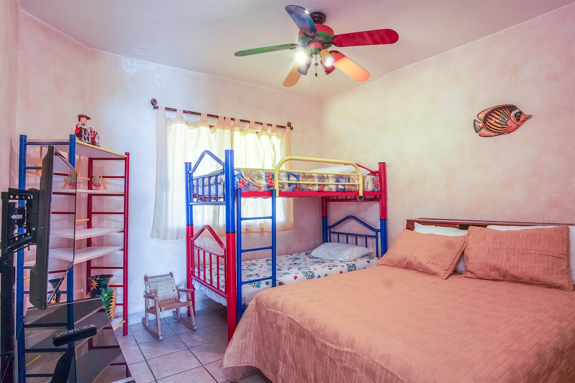 Habitación con literas, ventilador de techo, estantería, una silla pequeña y una cama con edredón rosa.