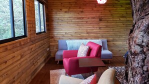 Interior acogedor de una cabaña con paredes de madera, un sofá azul, un sillón rojo y un gran tronco de árbol a la vista.