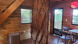 Interior de una cabaña de madera con un gran tronco de árbol, un refrigerador pequeño y una zona de comedor con sillas.