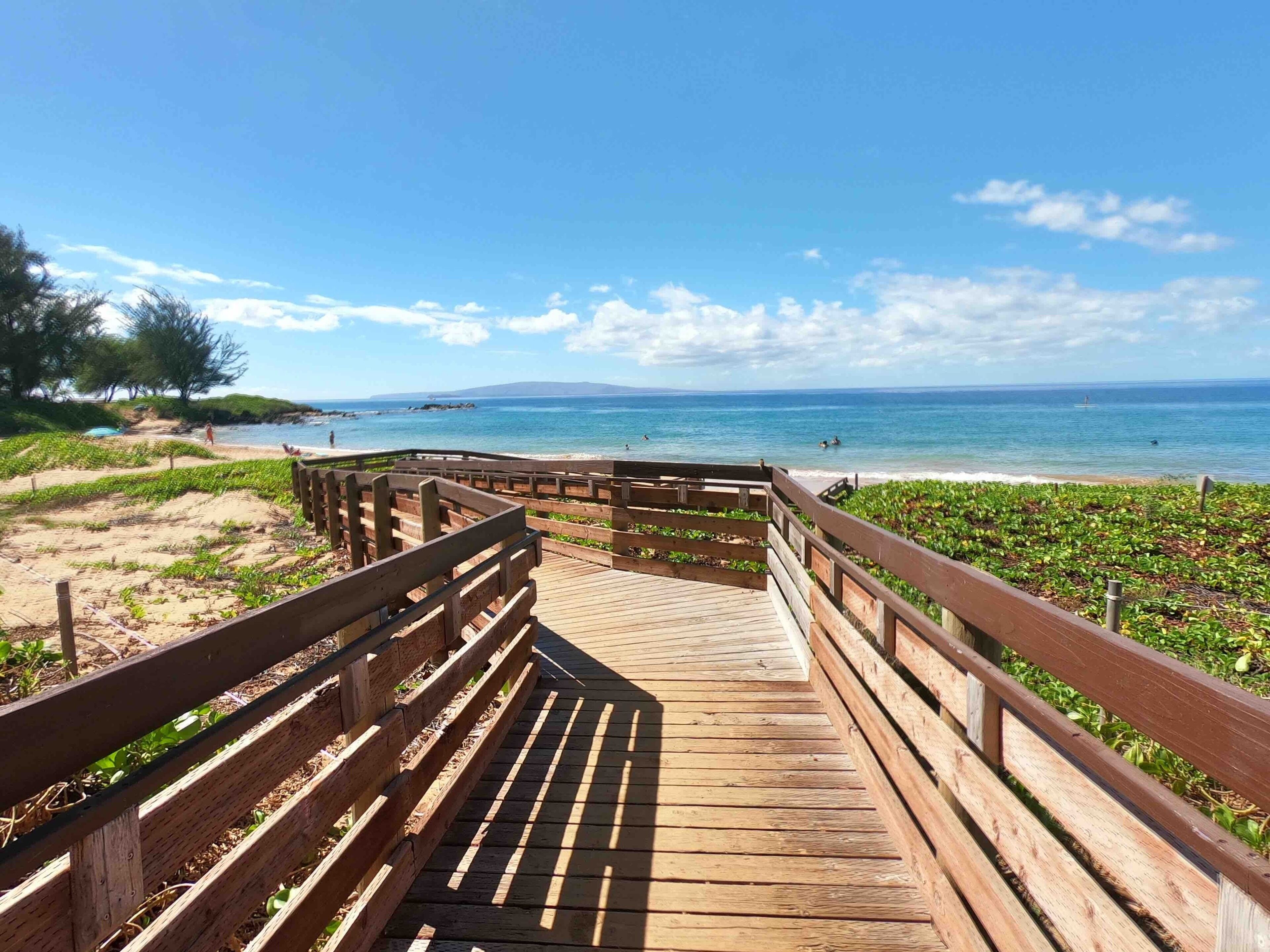 A wooden boardwalk leading to a beach with a clear view of the ocean and distant mountains.
