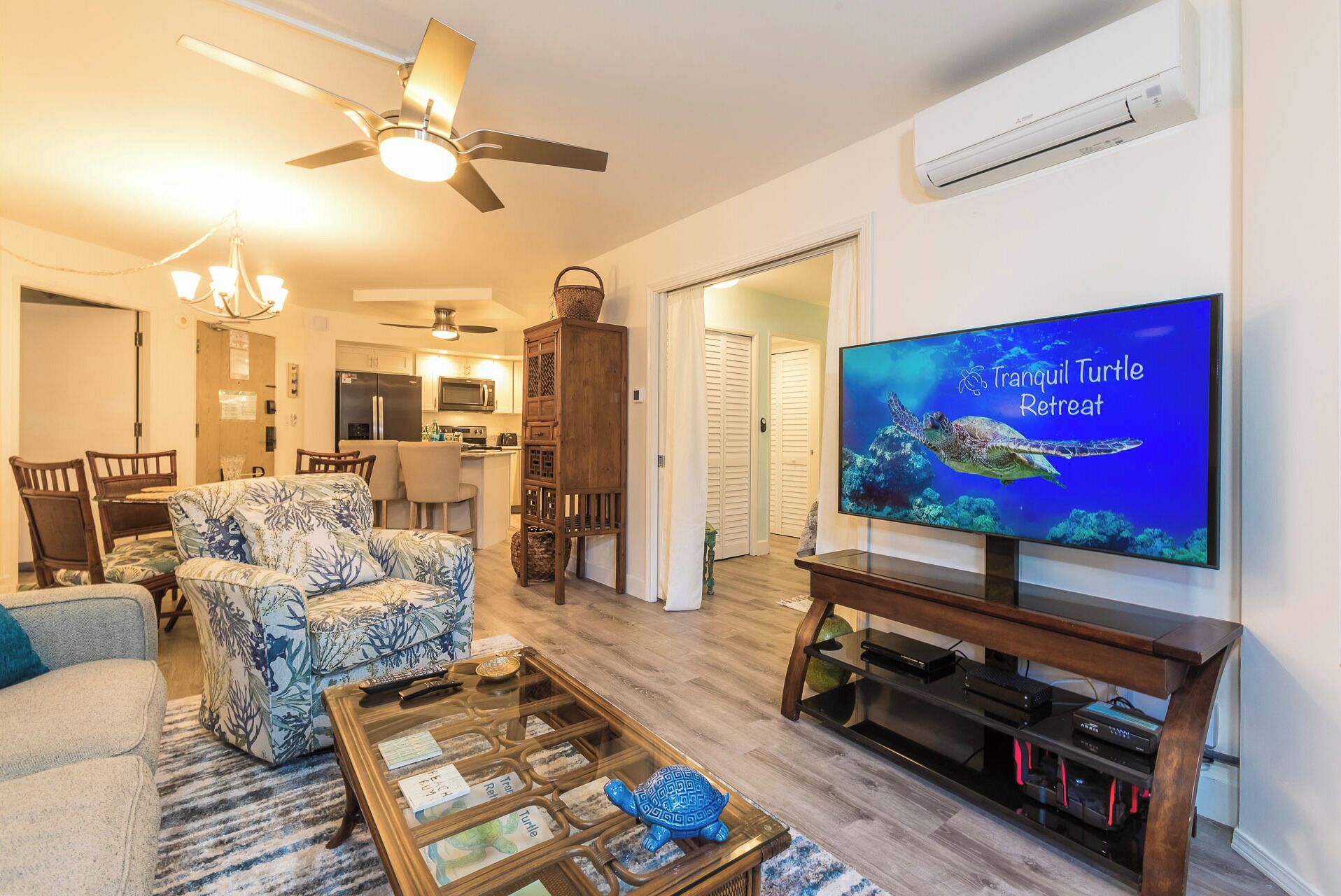 A living room with a television displaying 'Tranquil Turtle Retreat', a ceiling fan, a dining area with chairs, a kitchen, and a wooden cabinet.