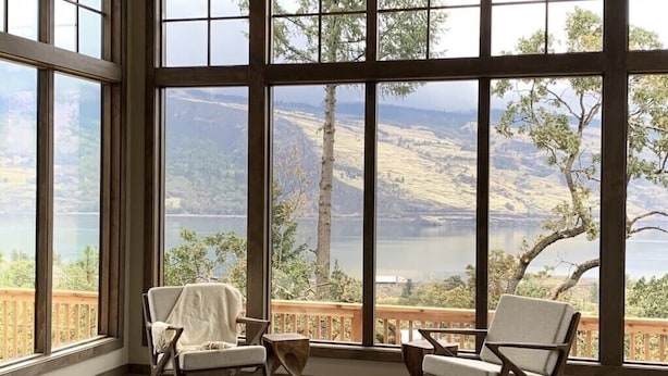 A room with large windows offering a view of a lake and hills. There are two chairs and a small table. - Newly built Mosier Bluff home-3 Bed, 3.5 Bath Spectacular River View and Hot Tub (Mosier)