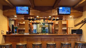 A bar area with wooden bar counter, four stools, two televisions mounted on the ceiling, and a variety of glassware and mugs displayed on the counter.