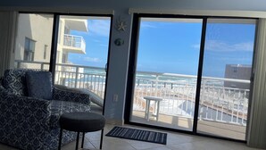 Un salón con sofá y mesa de centro, un libro, y vistas a la playa y al océano a través de grandes puertas correderas.