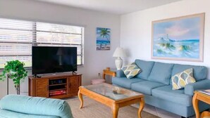 Una sala de estar con televisor, sofá, mesa de centro y un cuadro de una escena de playa.
