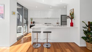 A modern kitchen with white cabinets, a central island with stools, and a large window. - Brand new house, amazing views, walk to the beach! (Kiama)