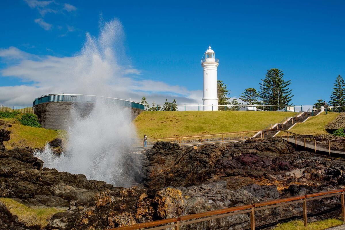 A lighthouse with a water spout, a stone structure, and a grassy area with steps leading up to the lighthouse.