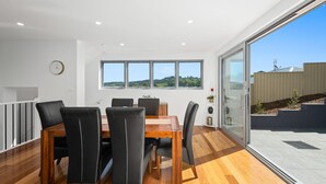 A dining area with a wooden table and black chairs, large windows with a view of greenery, and a glass door leading to an outdoor area. - Brand new house, amazing views, walk to the beach! (Kiama)